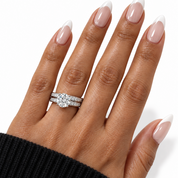 A close-up of a hand with a French manicure wearing a silver engagement ring with a round-cut center stone and pavé bands, alongside a matching pavé band ring, against a clean white background.