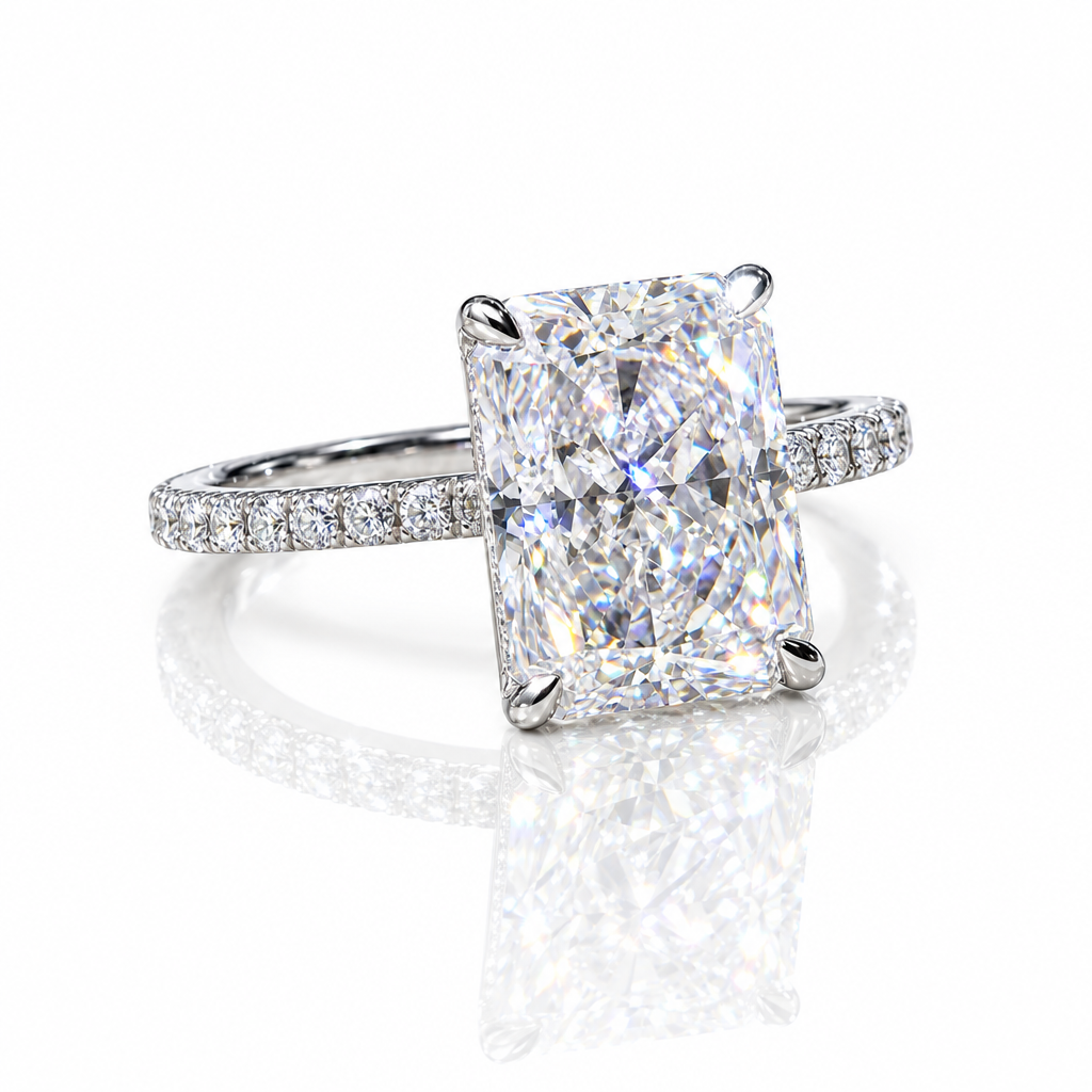 A radiant cut diamond engagement ring with a four-prong setting on a polished silver-colored band paved with small diamonds, shown on a reflective white surface.
