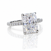 A radiant cut diamond engagement ring with a four-prong setting on a polished silver-colored band paved with small diamonds, shown on a reflective white surface.