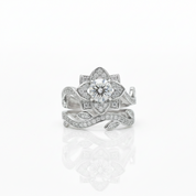 A silver-toned flower ring with a central round clear gemstone and surrounding pavé-set stones, shown on a clean white background with a soft reflection below.