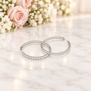 A pair of silver hoop earrings paved with clear stones, resting on a white marble surface with soft pink roses and white flowers blurred in the background.