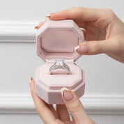 Hand holding a pink jewelry box with an engagement ring inside against a neutral background