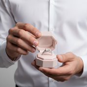 Person holding a pink jewelry box with an engagement ring inside, wearing a white shirt.