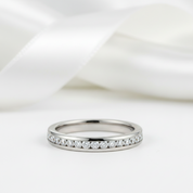 A slim silver eternity ring set with small round clear gemstones, photographed on a soft white surface with a blurred white satin ribbon in the background.