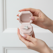 Hand holding a pink jewelry box with a ring inside against a light background