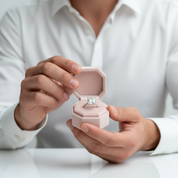 Person holding a pink jewelry box with a diamond ring inside, wearing a white shirt.