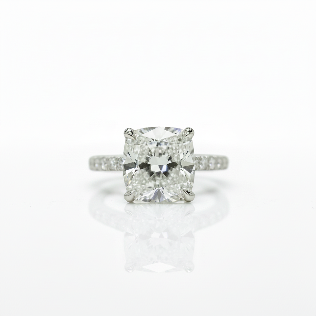 A radiant cut diamond engagement ring with a four-prong setting on a polished silver-colored band with small pavé diamonds, centered on a reflective white surface.