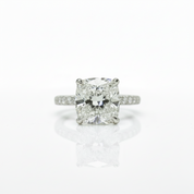 A radiant cut diamond engagement ring with a four-prong setting on a polished silver-colored band with small pavé diamonds, centered on a reflective white surface.
