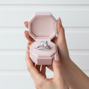 Hand holding a pink jewelry box with a diamond ring inside against a white background