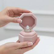 A close-up of hands holding an open pink octagonal jewelry box that displays a silver-toned ring set with a large clear center stone and smaller side stones.