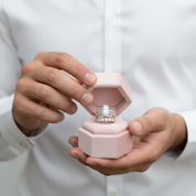 A close-up of a person in a white button-up shirt holding an open light pink jewelry box. The box contains a silver-toned ring with a large clear center stone and small clear side stones.