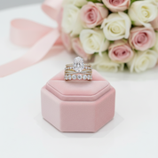 A three-band diamond ring set displayed on a pink velvet ring box, with a bouquet of pink and white roses in the soft-focus background.