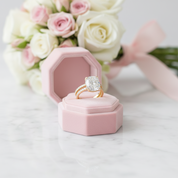 Gold ring with a diamond in a pink hexagonal box, surrounded by flowers on a marble surface.