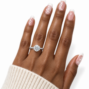 A close-up of a hand wearing a silver-toned diamond engagement ring with a round-cut center stone and a pavé band. The hand has a dark brown complexion with French manicure nails featuring white floral designs on a light pink base, and a cream knit sweater cuff is visible at the bottom.