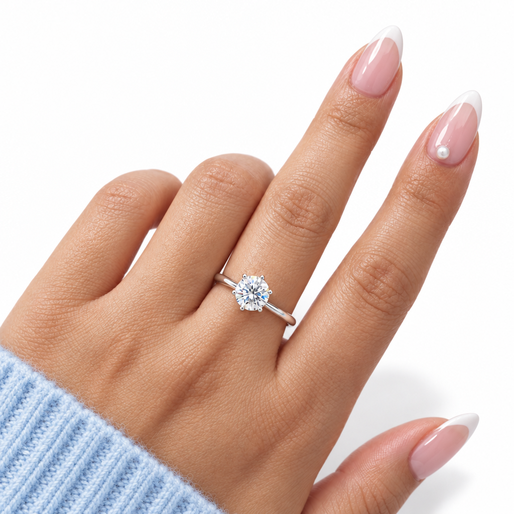 A close-up of a hand wearing a silver solitaire engagement ring with a round brilliant-cut diamond. The hand has a French manicure and is wearing a light blue knit sweater cuff, set against a clean white background.
