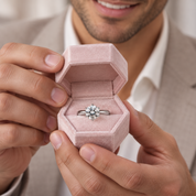 A close-up of a man holding an open dusty pink ring box that displays a silver-toned ring with a round brilliant-cut diamond. The man is wearing a light-colored collared shirt and a beige blazer, with a soft-focus background.