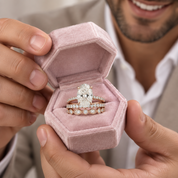A close-up lifestyle shot of a man holding an open dusty pink velvet ring box, showcasing a diamond engagement ring and matching wedding band set.
