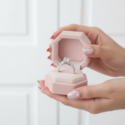 Hand holding a pink jewelry box with a silver ring inside against a white background