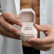 Person holding a pink jewelry box with an engagement ring inside, wearing a white shirt.