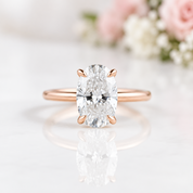 A rose gold engagement ring with a large oval-cut diamond set in a four-prong setting, resting on a white marble surface with a soft reflection and blurred pink flowers in the background.