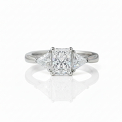 A classic three-stone diamond ring featuring a princess-cut center stone flanked by two smaller round diamonds, set in a polished silver-colored band on a reflective white surface.