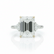An emerald-cut diamond ring with triangular side diamonds on a silver-toned band with gold accents, centered on a white background with a soft reflection.