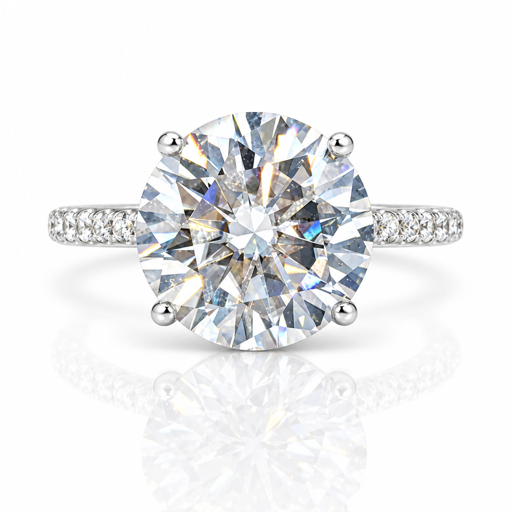 A round brilliant-cut diamond solitaire engagement ring with a silver-toned four-prong setting and pavé diamond accents on the band, shown on a clean white background with a soft reflection below.