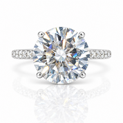 A round brilliant-cut diamond solitaire engagement ring with a silver-toned four-prong setting and pavé diamond accents on the band, shown on a clean white background with a soft reflection below.