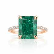 An emerald-cut green gemstone ring with a rose gold band accented by small pavé diamonds, shown on a clean white background with a soft reflection.