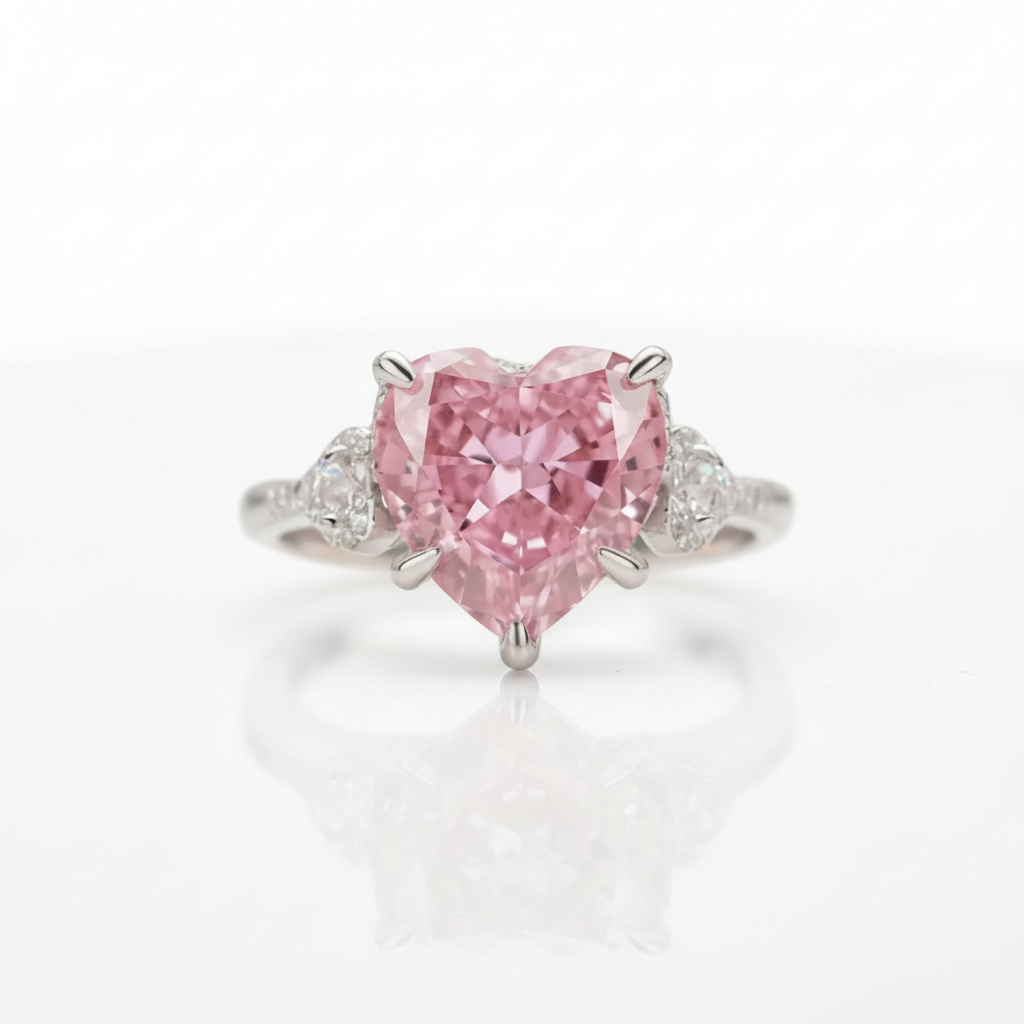 A heart-shaped pink diamond ring set in a silver-colored metal band with small round diamonds on either side, photographed on a clean white reflective surface.