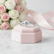 A silver-toned diamond ring with a central round-cut stone and twisted side details, displayed on a small pink velvet ring box. Pink roses and a pale pink ribbon are softly blurred in the background on a light marble surface.