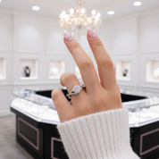 A close-up of a hand wearing a silver ring with a clear round-cut gemstone, set against a softly blurred jewelry store background with display cases and a chandelier.