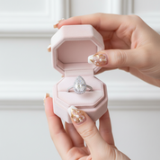 Hand holding a pink jewelry box with a diamond ring inside against a white background