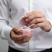 Person holding a pink jewelry box with a diamond ring inside, wearing a white shirt.