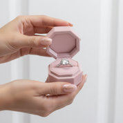 Hand holding a pink jewelry box with an engagement ring inside against a white background