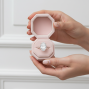 Hand holding a pink jewelry box with an engagement ring inside against a white background
