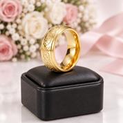 A gold ring with intricate floral engraving sits on a black square jewelry box, with blurred pink roses, white filler flowers, and a pink ribbon in the background.
