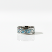 A polished silver-toned band ring featuring an intricate inlay of light blue, white, and gray floral and geometric patterns, displayed on a clean white reflective surface.