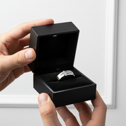 A silver band ring with engraved lettering sits inside an open black jewelry box, held by two hands against a soft white background.