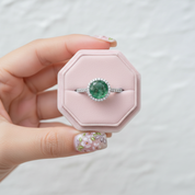A hand with floral nail art holds a light pink octagonal ring box containing a silver ring with a round green gemstone surrounded by small clear stones, against a soft white textured background.