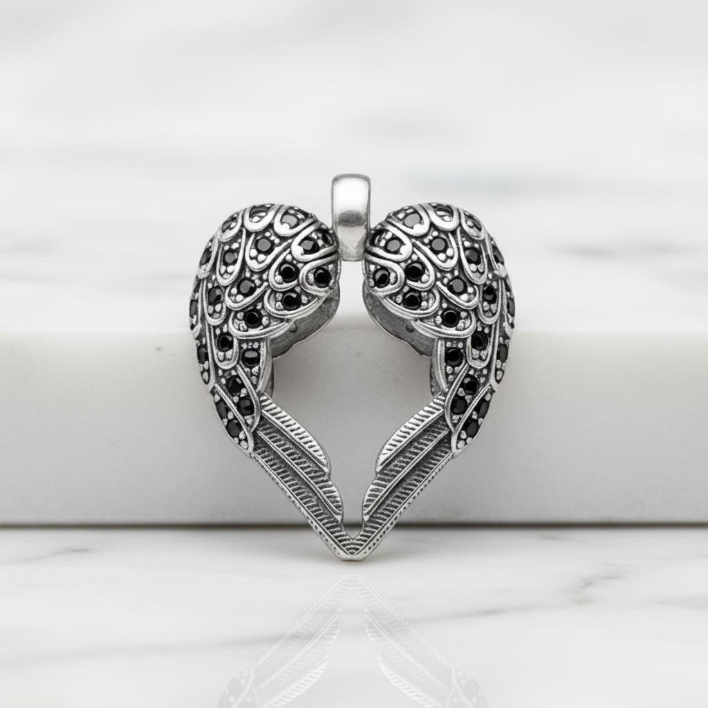 A silver-toned heart-shaped pendant with intricate filigree wing details and small dark stones, displayed on a white marble surface.