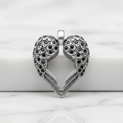 A silver-toned heart-shaped pendant with intricate filigree wing details and small dark stones, displayed on a white marble surface.