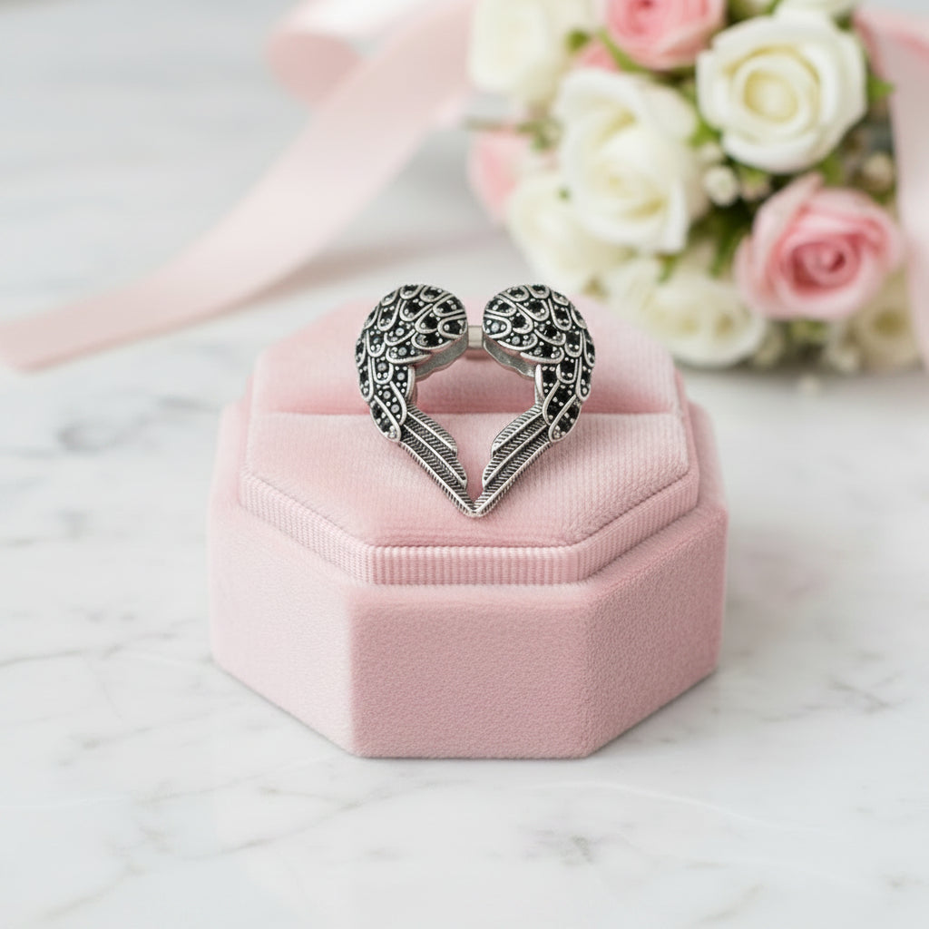 A silver heart-shaped ring with intricate filigree detailing, displayed on a pink velvet hexagonal ring box with a soft pink ribbon and blurred pink and white roses in the background.