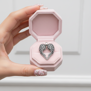 A hand with decorated nails holds an open light pink jewelry box containing a silver heart-shaped pendant with intricate filigree wings.