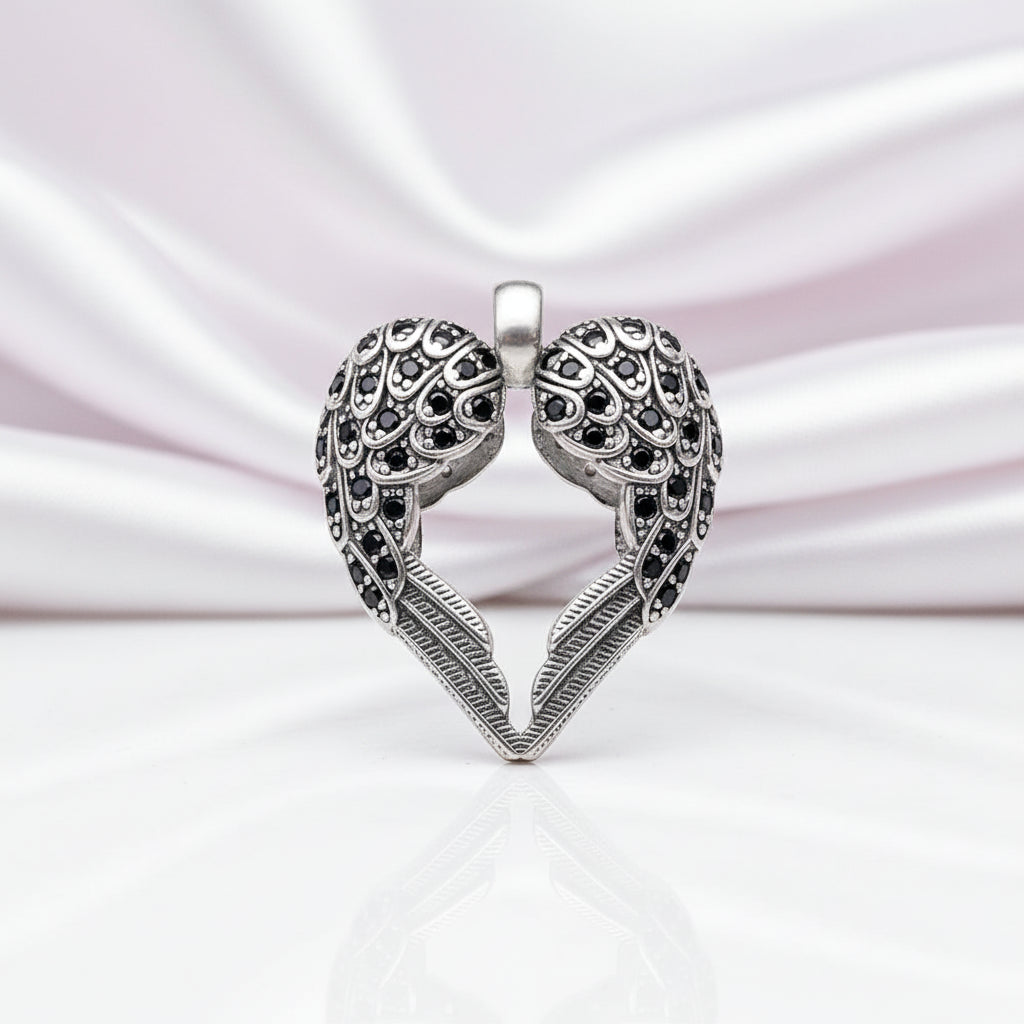 A silver heart-shaped pendant with two angel wings, embellished with small black stones, displayed on a reflective white surface against a soft pink satin background.