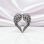 A silver heart-shaped pendant with two angel wings, embellished with small black stones, displayed on a reflective white surface against a soft pink satin background.