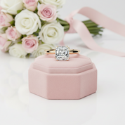 A square emerald-cut diamond ring with a yellow gold band, displayed on a pink hexagonal ring box with a bouquet of pink and white roses in the background.