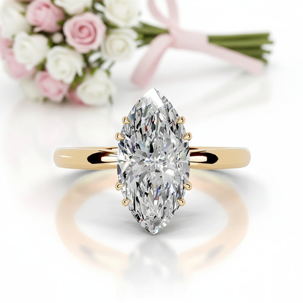 A marquise-cut diamond engagement ring with a yellow gold band, shown in a clean studio setting with a soft reflection below and a blurred bouquet of pink and white roses in the background.