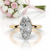 A marquise-cut diamond engagement ring with a yellow gold band, shown in a clean studio setting with a soft reflection below and a blurred bouquet of pink and white roses in the background.