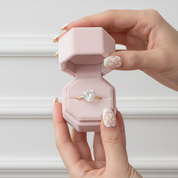 A heart-shaped diamond ring on a gold band, presented inside an open light pink velvet ring box held by two hands with floral nail art.